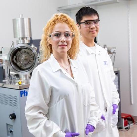 Two individuals in white lab coats and goggles standing in front of machinery 